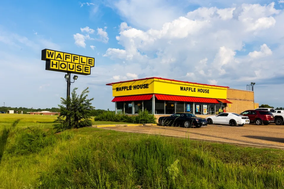 Waffle House restaurant in rural open area with large yellow and black sign and several cars parked in front.